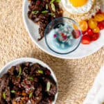 Beef tapa in a bowl with fork and a plate of garlic rice, sunny side egg, tomato, spiced vinegar, and tapa at the top right.