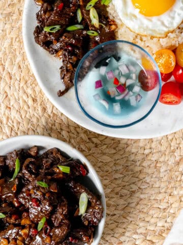 Beef tapa in a bowl with fork and a plate of garlic rice, sunny side egg, tomato, spiced vinegar, and tapa at the top right.